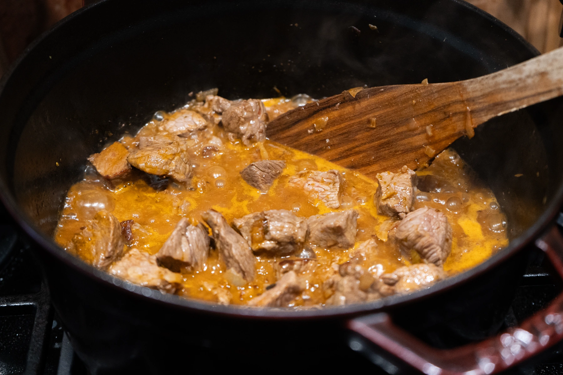 Fleischstücke in brauner Soße blubbern in einem Topf. Ein Holzlöffel rührt die Mischung um.