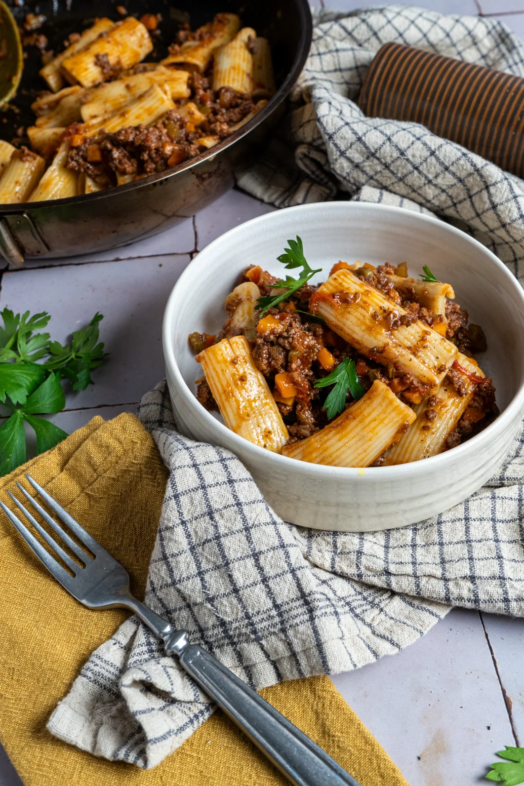 Weiße Schüssel mit Rigatoni und Hackfleischsauce, garniert mit Petersilie. Besteck und Servietten neben der Schüssel.