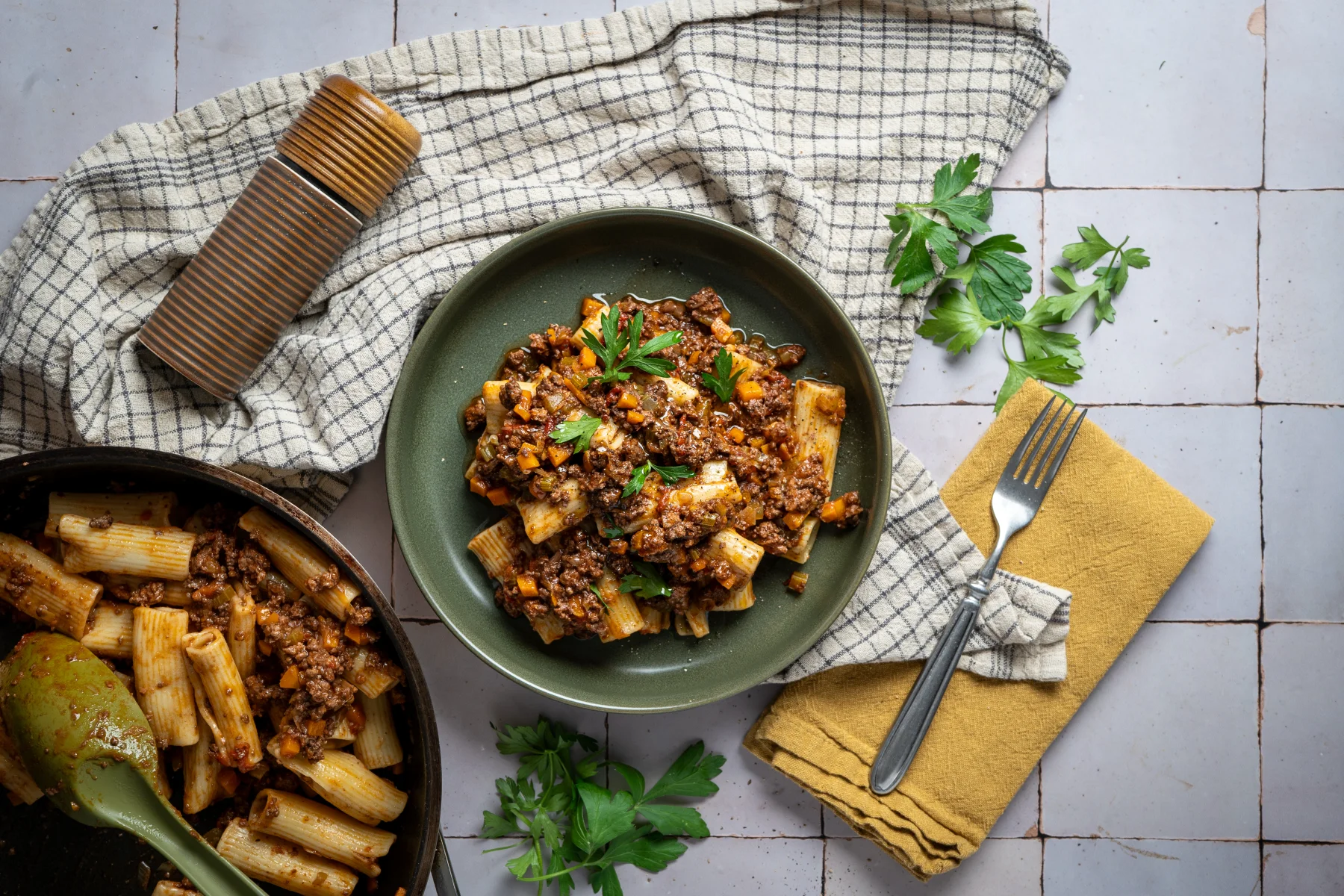 Grüner Teller mit Pasta Bolognese, dekoriert mit Petersilie, daneben eine Gabel auf Serviette und ein Gewürzstreuer.