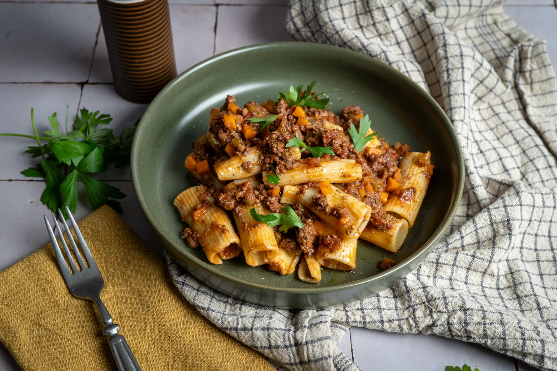 Grüner Teller mit Rigatoni, Hackfleischsauce und Petersilie, daneben Besteck auf einer gelben Serviette und ein Küchentuch.