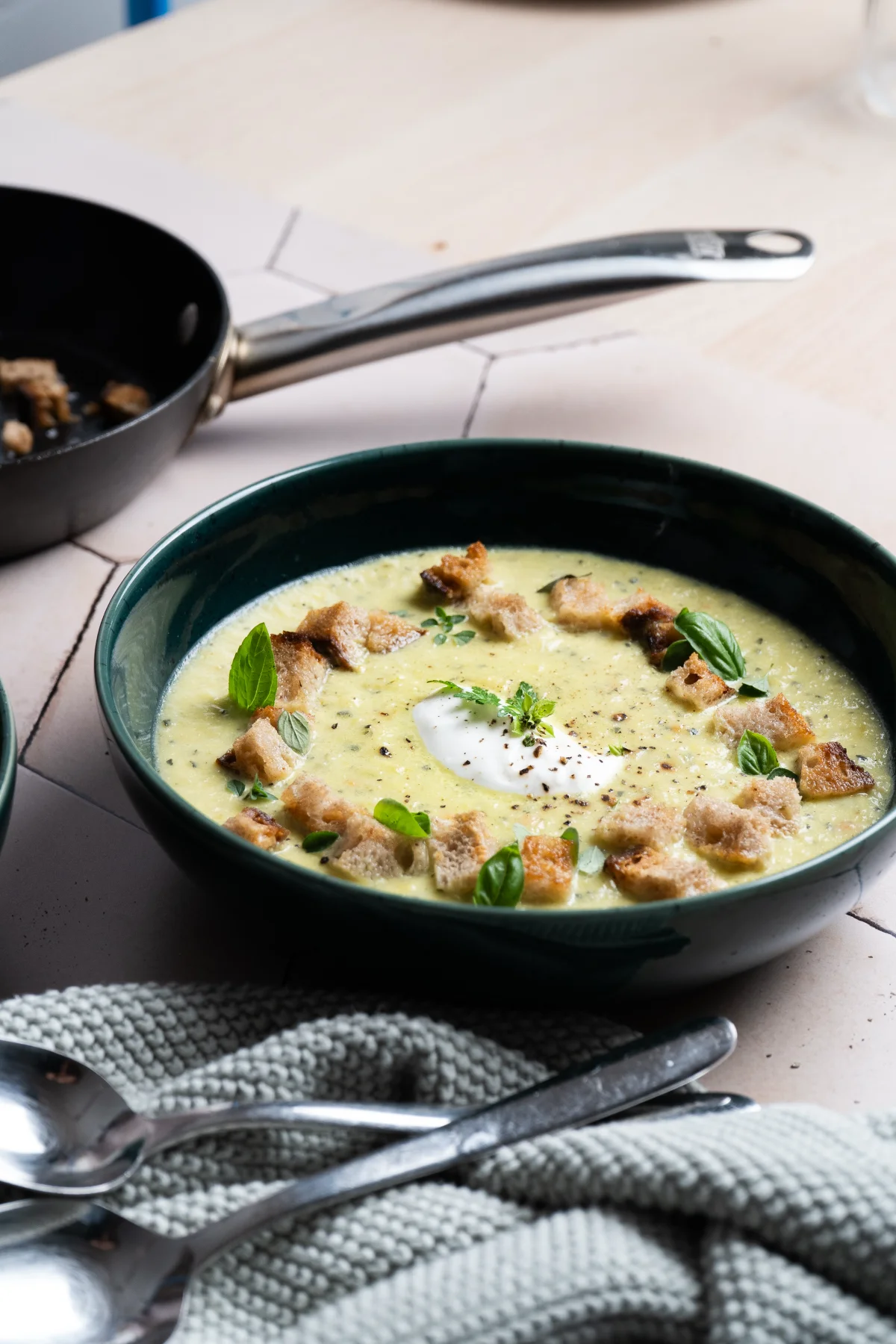 Grüne Suppe in einer Schüssel, garniert mit Croutons, Basilikum und einem Klecks Sahne. Im Hintergrund eine Pfanne.