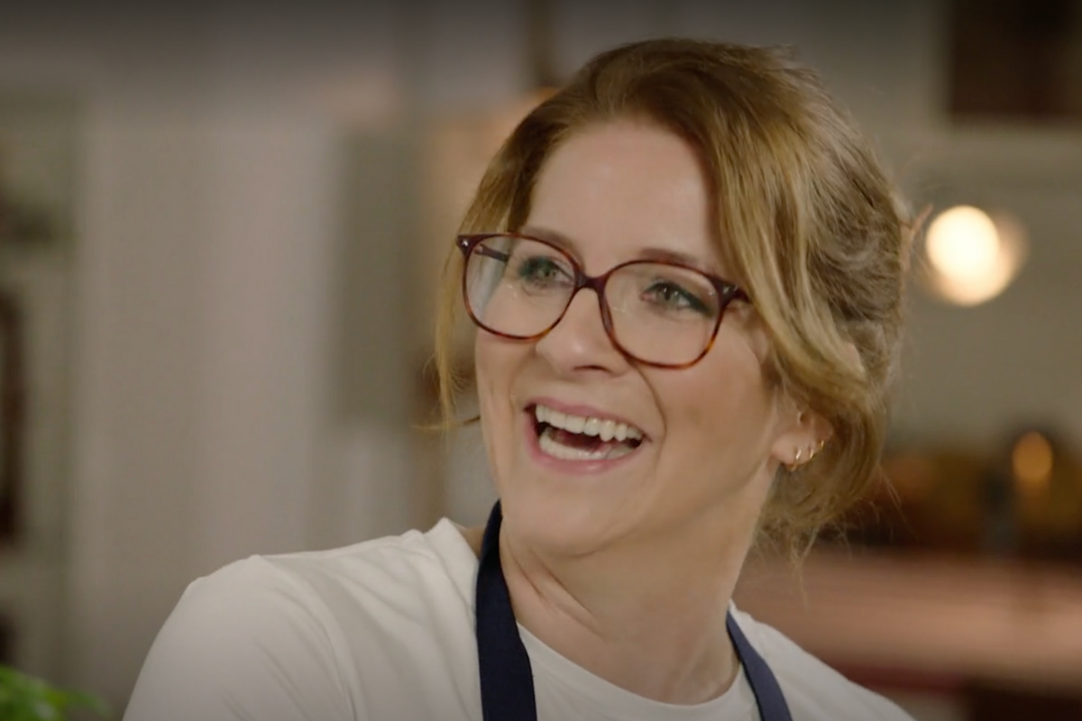 einfach-&-köstlich-3 Lachende Frau mit Brille und hochgestecktem Haar. Sie trägt ein weißes Oberteil und eine Schürze. Hintergrund unscharf.