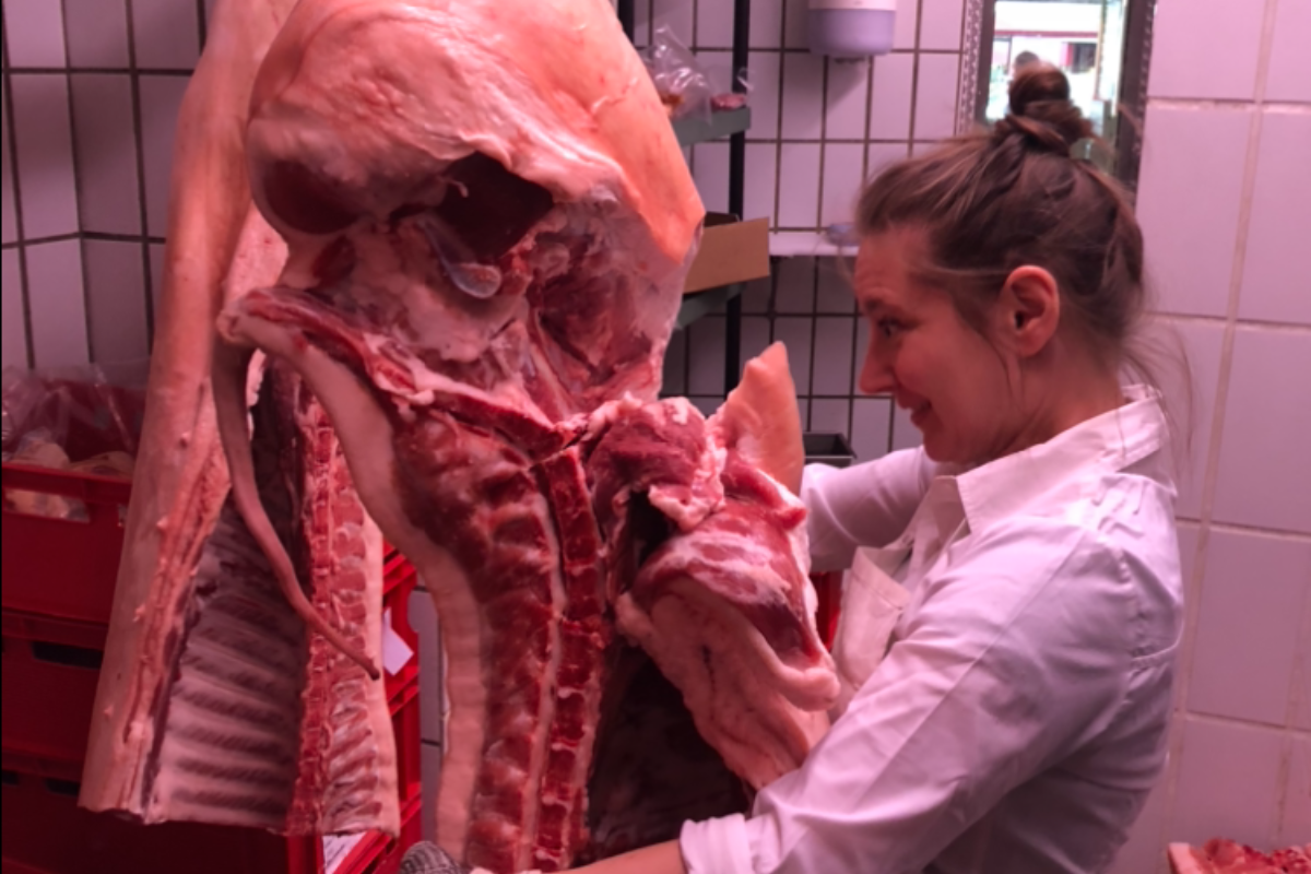 Eine Person in weißem Kittel betrachtet ein großes Stück Fleisch in einem Kühlraum mit weißen Fliesen.