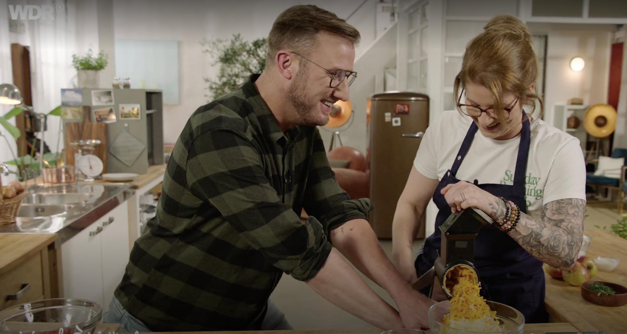 einfach-&-köstlich-1 Ein Mann und eine Frau reiben gemeinsam Gemüse in einer modernen Küche. Beide lächeln und genießen die Tätigkeit.