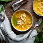Zwei Schalen klassische Hühnersuppe mit Hühnerfleisch, Karotten und Sellerie, daneben eine Suppenkelle, Pfeffermühle und Lorbeerblätter auf einem grauen Tuch.
