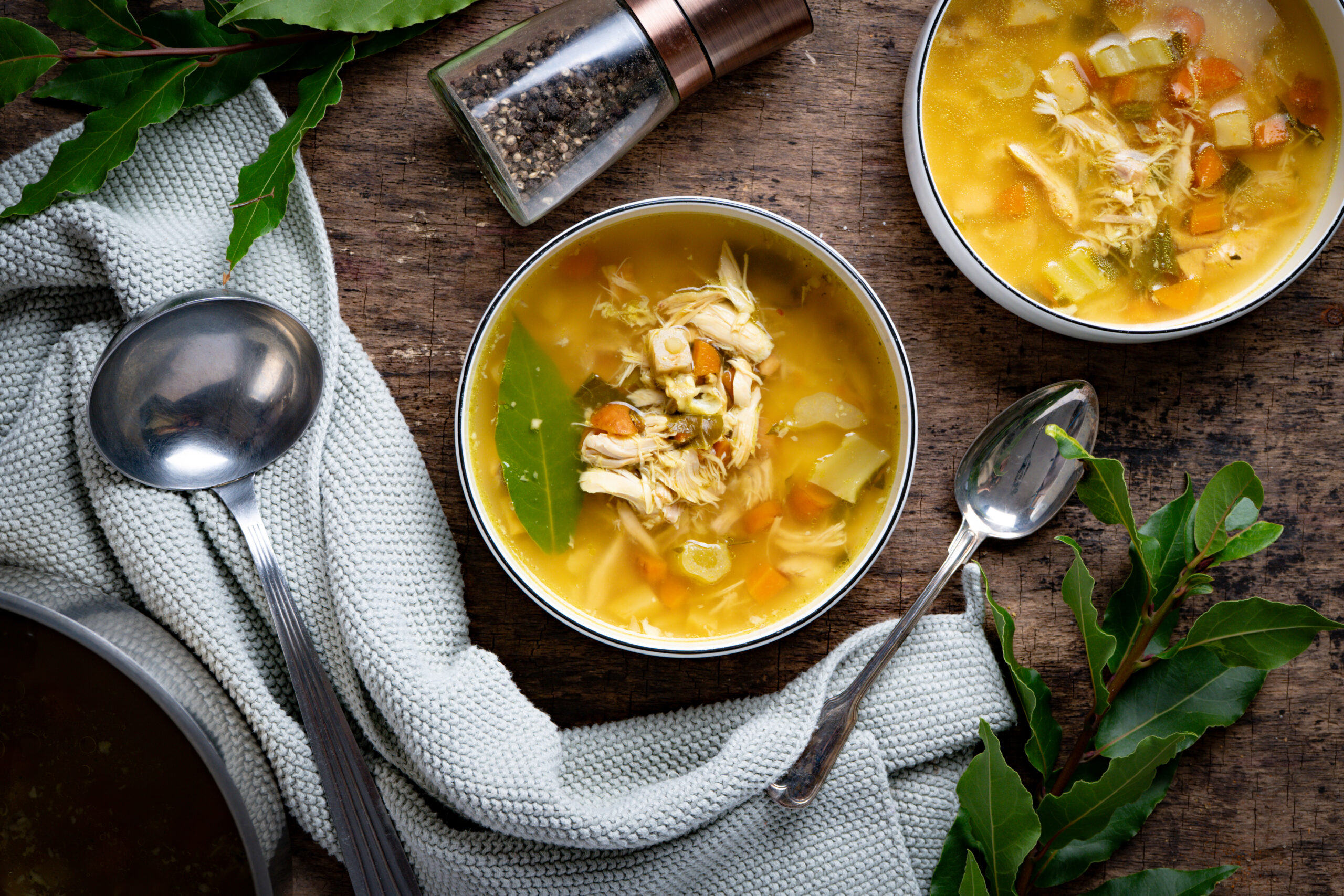 Klassische Hühnersuppe Zwei Schalen klassische Hühnersuppe mit Hühnerfleisch, Karotten und Sellerie, daneben eine Suppenkelle, Pfeffermühle und Lorbeerblätter auf einem grauen Tuch.