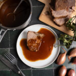 Ein Teller mit Bratenscheibe in Soße, daneben ein Topf mit Soße, Schalotten und aufgeschnittener Braten auf Holzbrett.