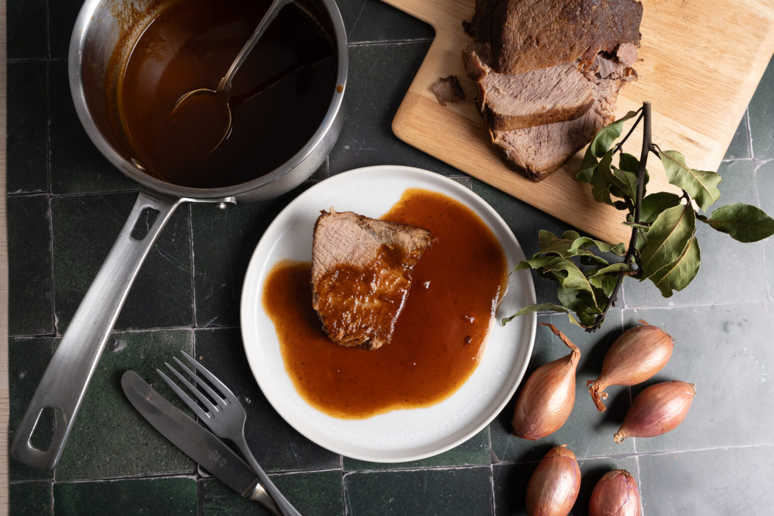 Ein Teller mit Bratenscheibe in Soße, daneben ein Topf mit Soße, Schalotten und aufgeschnittener Braten auf Holzbrett.