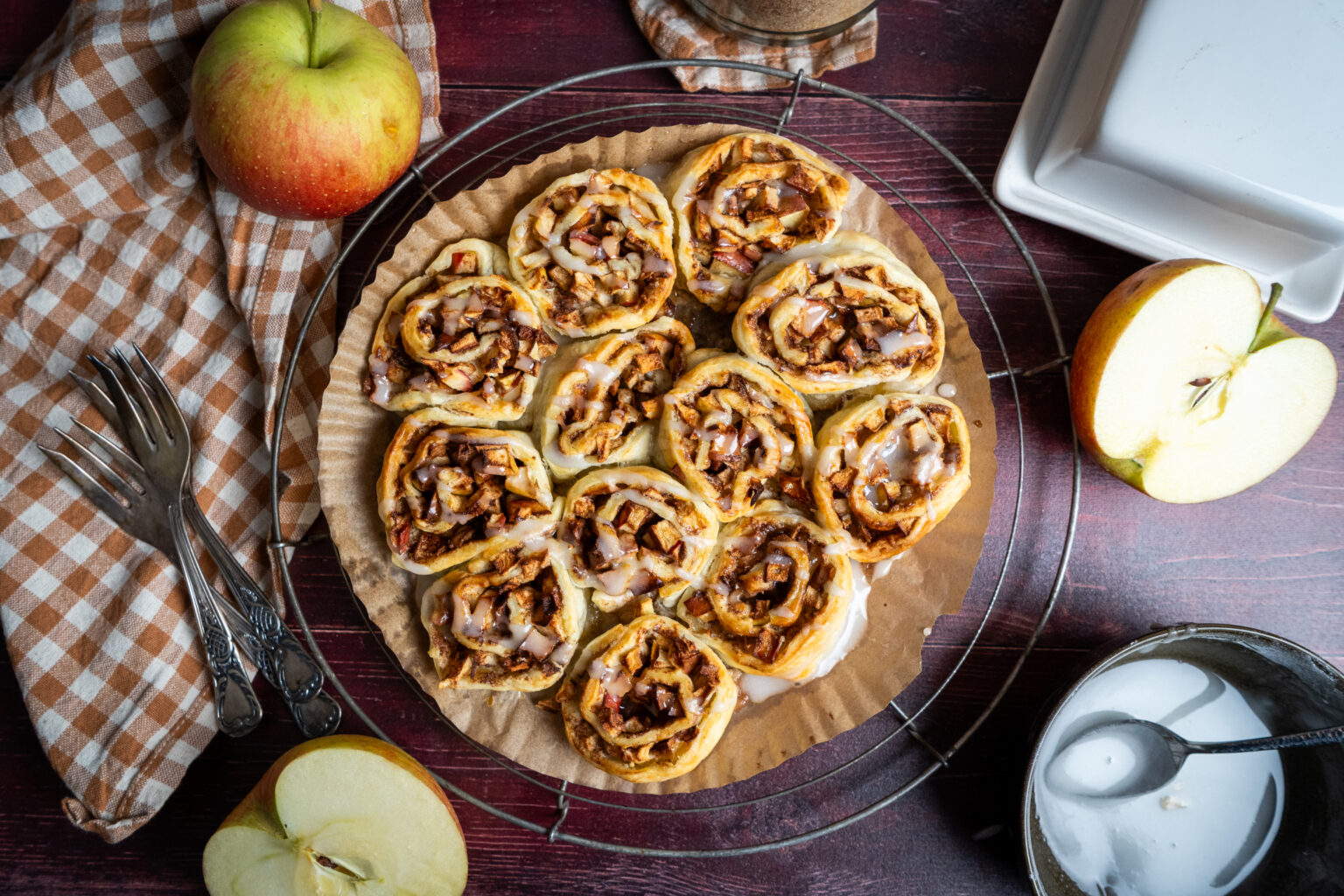 Frisch gebackene Apfelschnecken mit Zuckerguss auf einem Gitter, daneben eine aufgeschnittene und eine ganze Apfel.