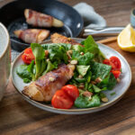 Teller mit Feldsalat, Cherrytomaten, gehackten Mandeln und einer Birne im Speckmantel, im Hintergrund eine Pfanne mit weiteren Speckbirnen und Zitrone.