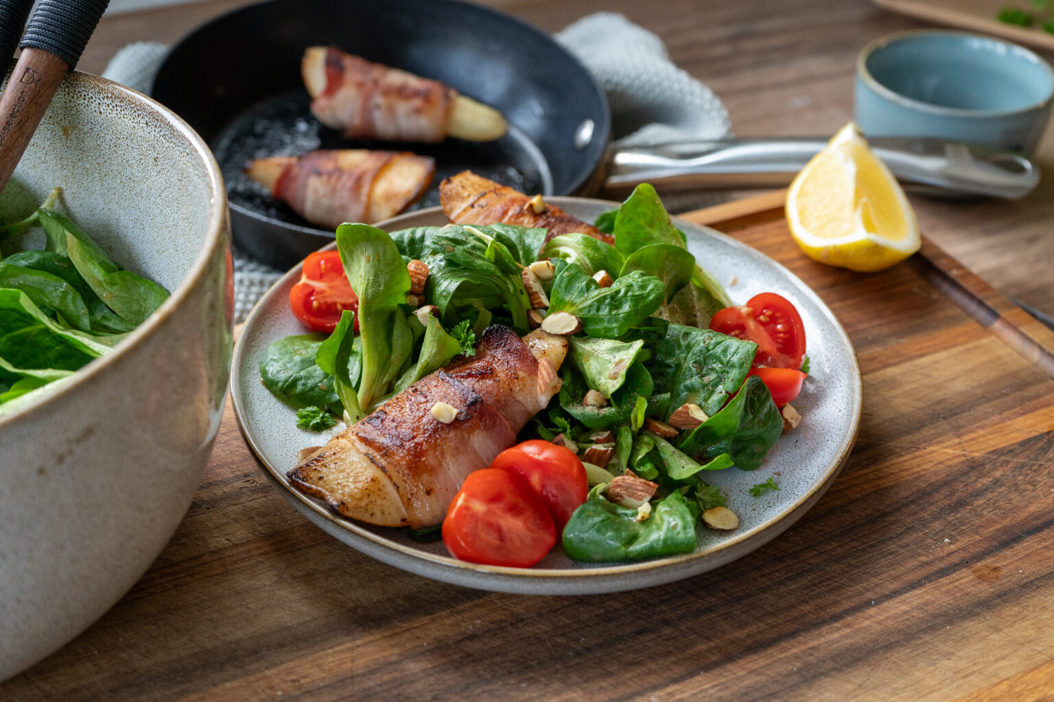 Teller mit Feldsalat, Cherrytomaten, gehackten Mandeln und einer Birne im Speckmantel, im Hintergrund eine Pfanne mit weiteren Speckbirnen und Zitrone.