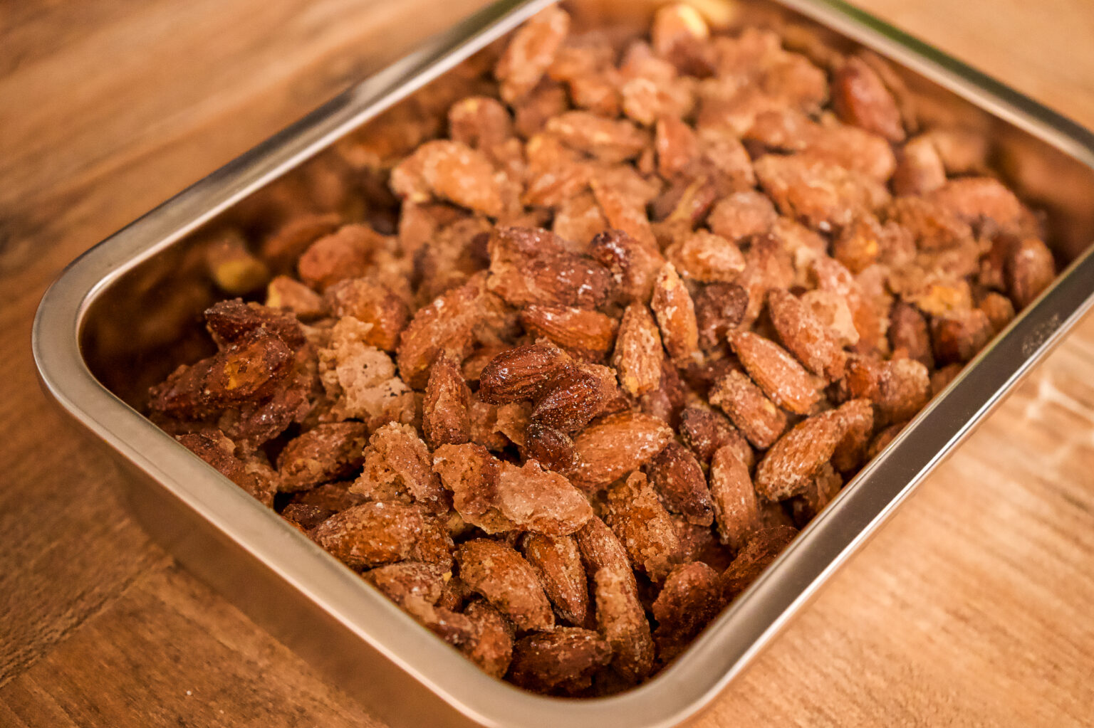 Metallschale mit frisch gebrannten Mandeln auf einem Holztisch. Die Mandeln sind knusprig und mit Zucker überzogen.