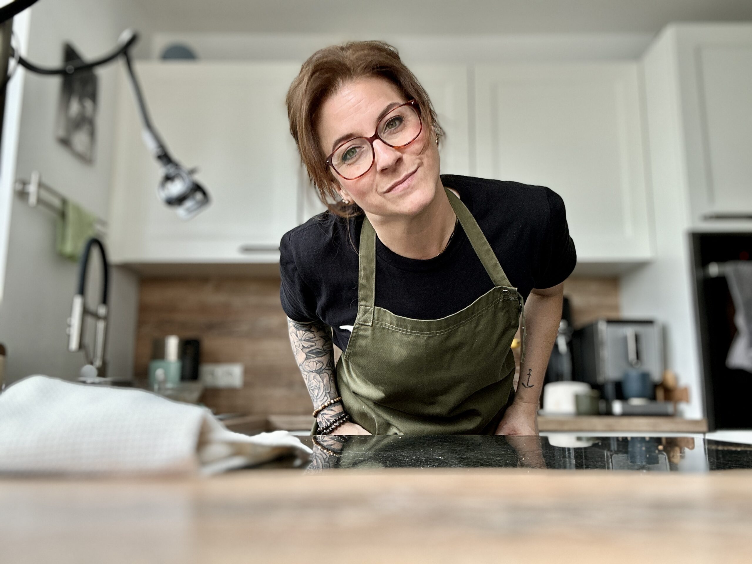 Person in grüner Schürze lehnt sich über Küchenarbeitsfläche, lächelt in die Kamera. Küche im Hintergrund.