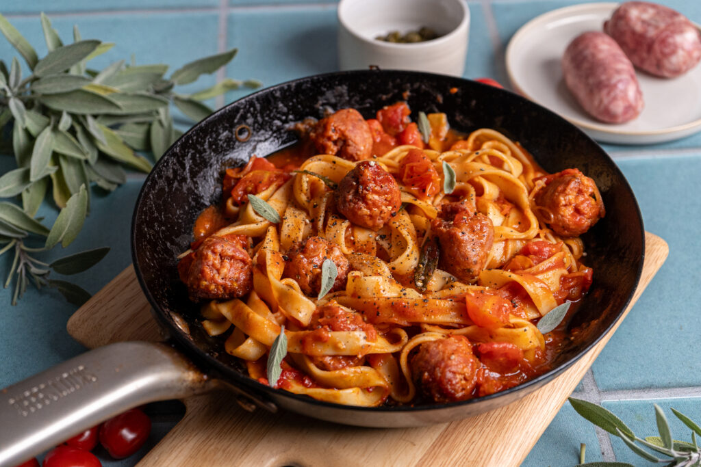 Pfanne mit Tagliatelle, Tomatensauce und Fleischbällchen, garniert mit frischem Salbei, auf einem Holzbrett.