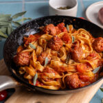 Pfanne mit Tagliatelle, Tomatensauce und Fleischbällchen, garniert mit frischem Salbei, auf einem Holzbrett.