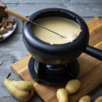 Fondue-Topf mit geschmolzenem Käse, Gabel, daneben Brotstücken, Kartoffeln und Birne auf Holztisch und Brett.