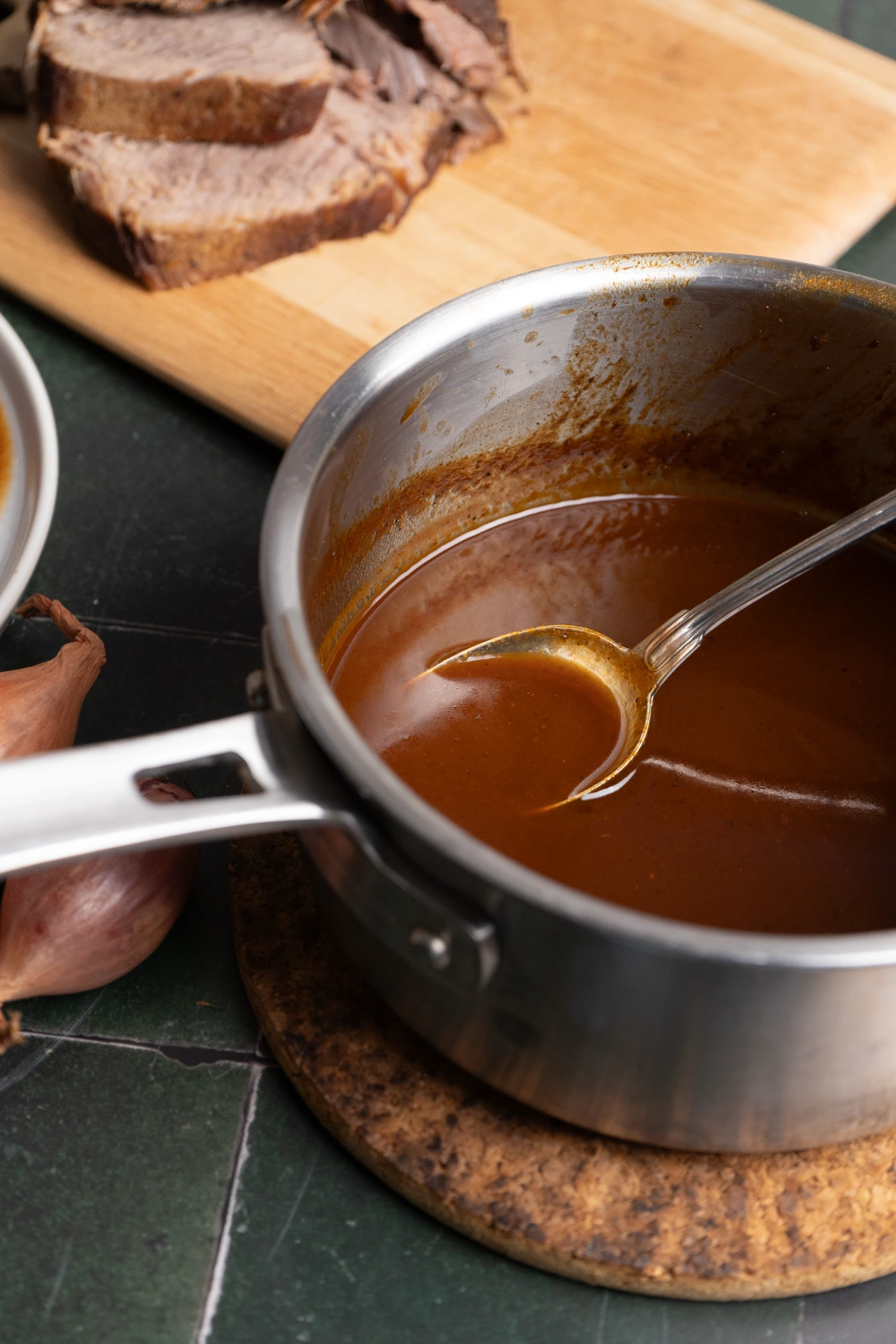 Edelstahltopf mit brauner Sauce und Löffel, dahinter geschnittenes Fleisch auf Holzbrett.