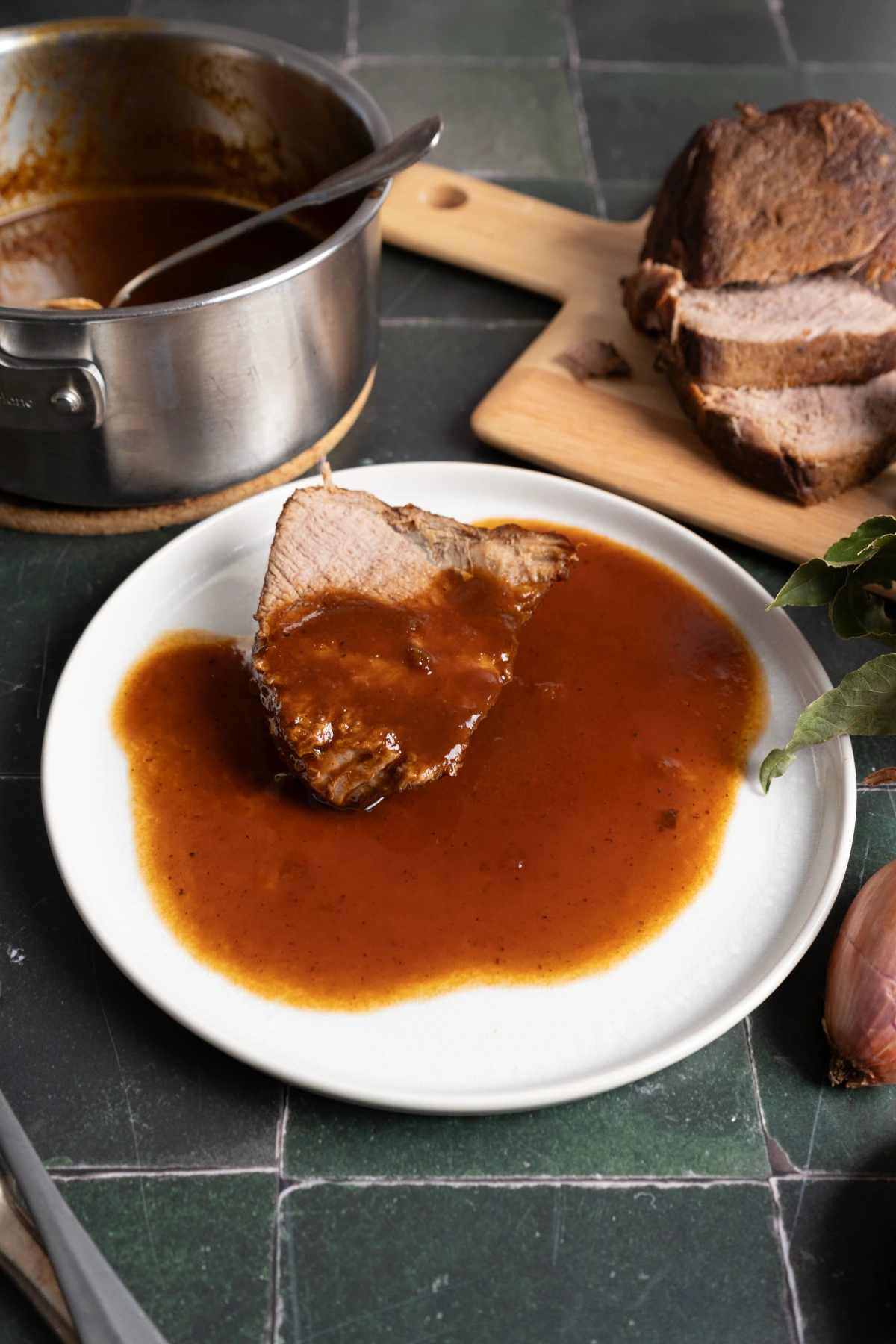 Eine Scheibe Braten mit brauner Soße auf einem weißen Teller, daneben ein Topf und geschnittener Braten auf einem Holzbrett.