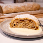 Stück Apfelstrudel mit Puderzucker auf einem Teller mit Vanillesauce, dahinter der restliche Strudel auf einem Holzbrett.