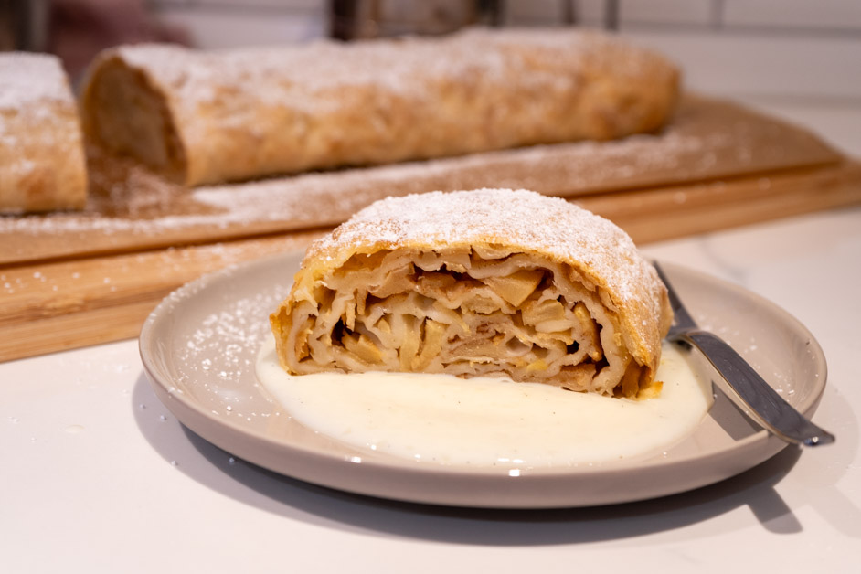 Apfelstrudel mit Vanillesauce Stück Apfelstrudel mit Puderzucker auf einem Teller mit Vanillesauce, dahinter der restliche Strudel auf einem Holzbrett.