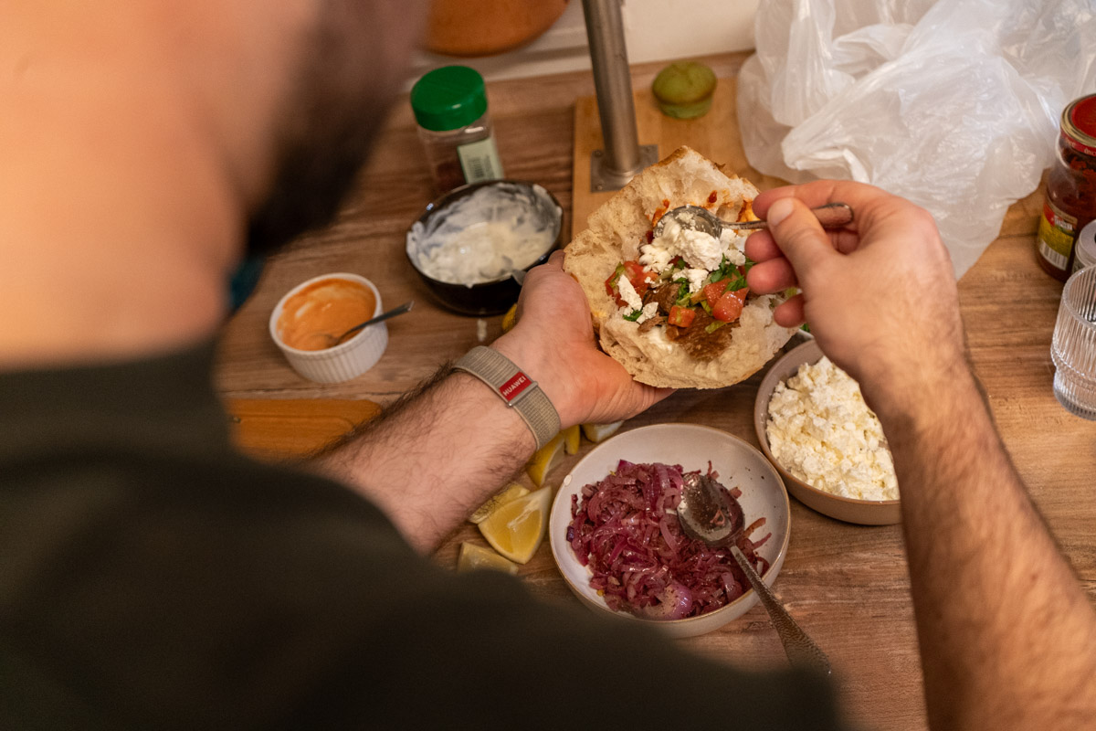 Person füllt Fladenbrot mit Gemüse und Käse, umgeben von verschiedenen Zutaten in Schüsseln auf einer Küchenarbeitsfläche.