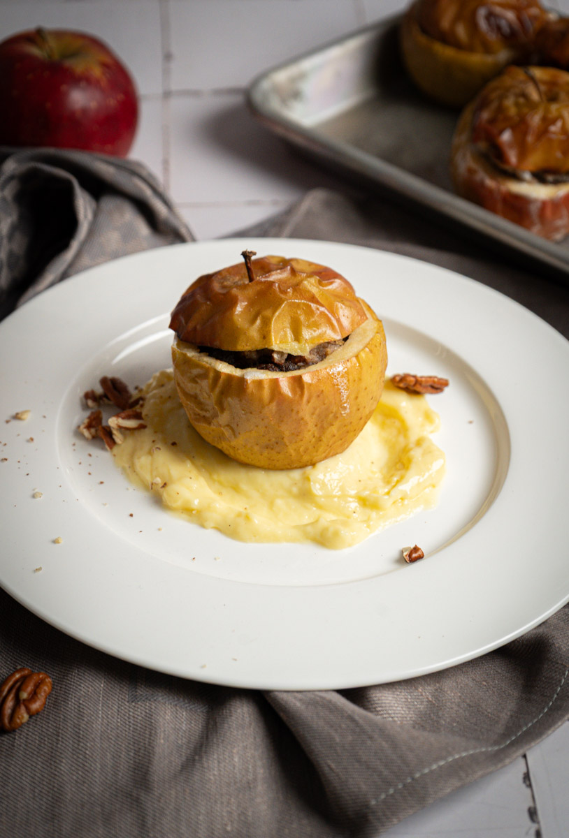 Gebackener Apfel auf Vanillesauce, garniert mit Nüssen, auf einem weißen Teller serviert. Im Hintergrund ein roter Apfel.