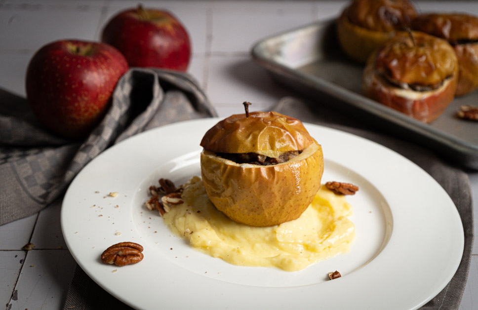 Gebackener Apfel auf Vanillesauce, garniert mit Pekannüssen, im Vordergrund; zwei frische Äpfel und Backblech im Hintergrund.