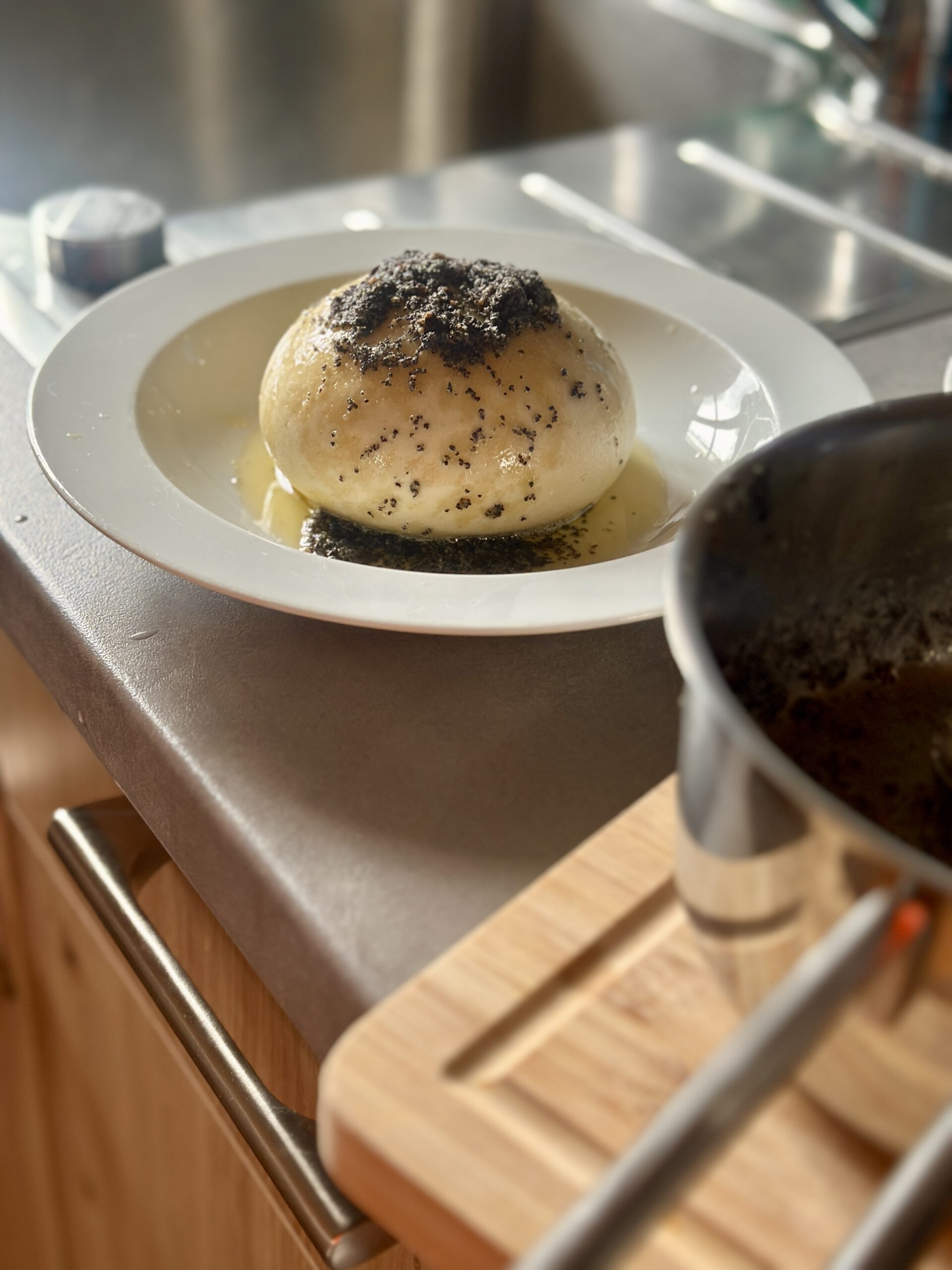 Germknödel mit Zwetschgenmus Gedämpfter Knödel mit Mohn und Butter auf einem weißen Teller, neben einem Topf in einer Küche.