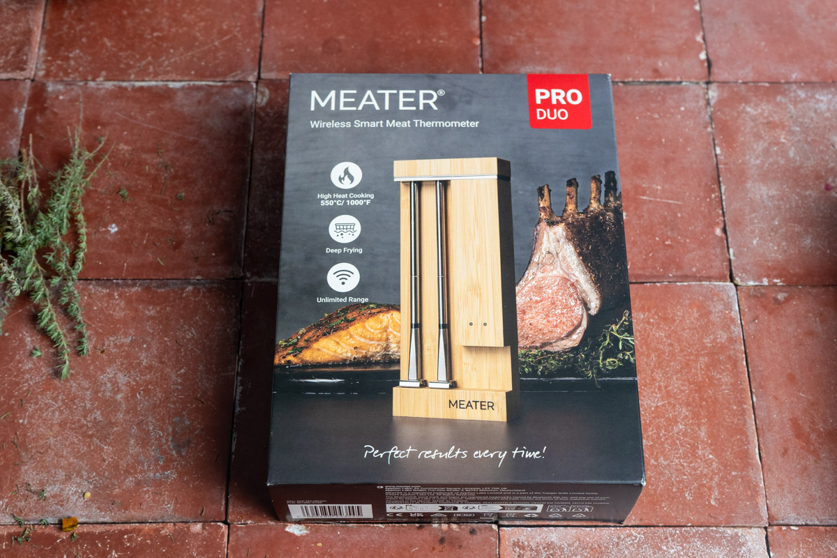 Verpackung eines kabellosen Fleischthermometers auf Fliesenboden, daneben etwas Kräuter.