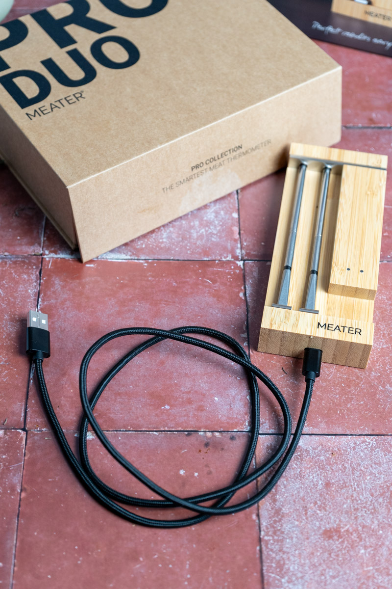 Holz-Ladestation mit zwei Metallsonden und USB-Kabel auf rotem Fliesenboden. Kartonverpackung daneben.