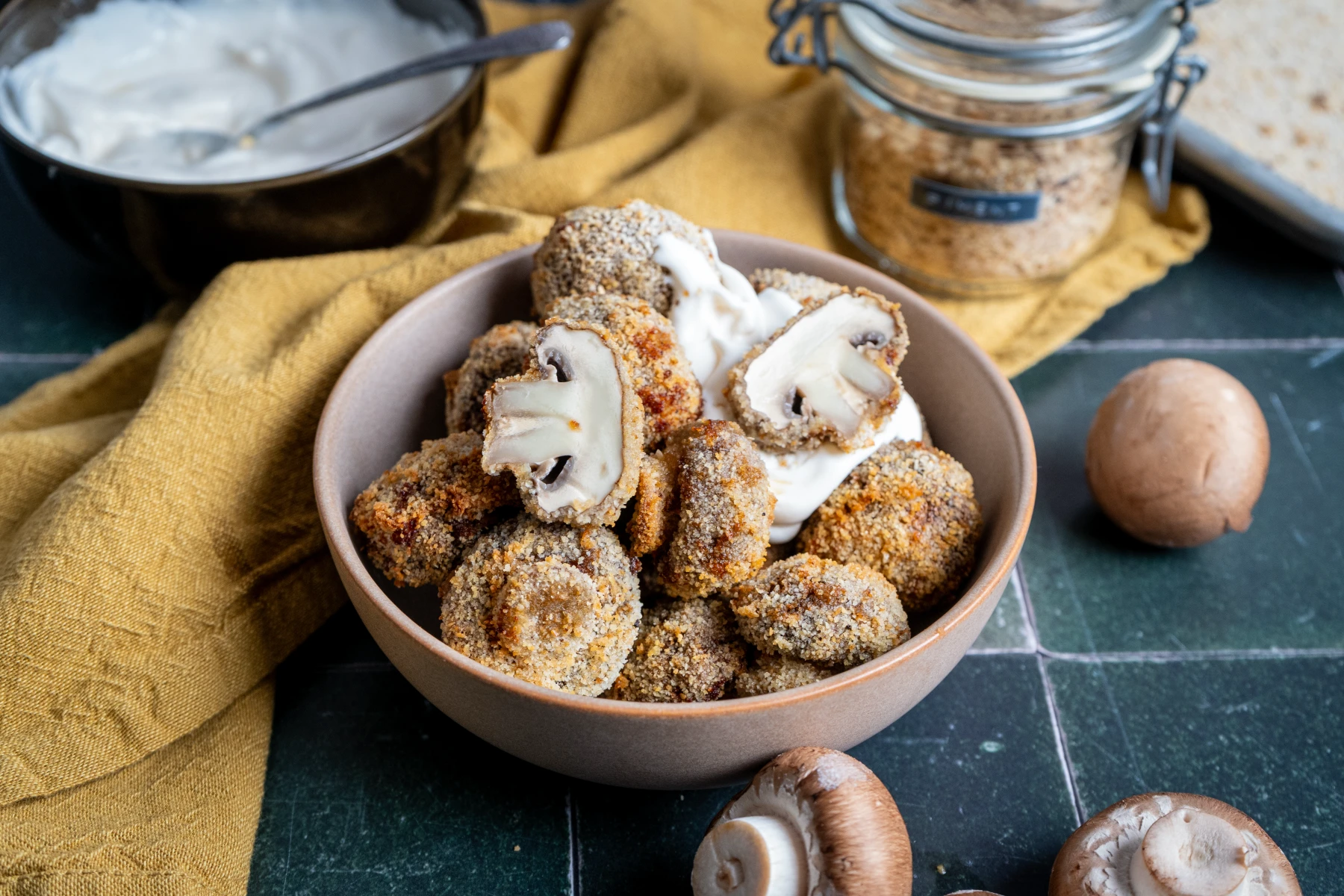 Gebackene, panierte Champignons in einer Schüssel, daneben ein Dip und einige ungekochte Pilze auf einem Holzbrett.