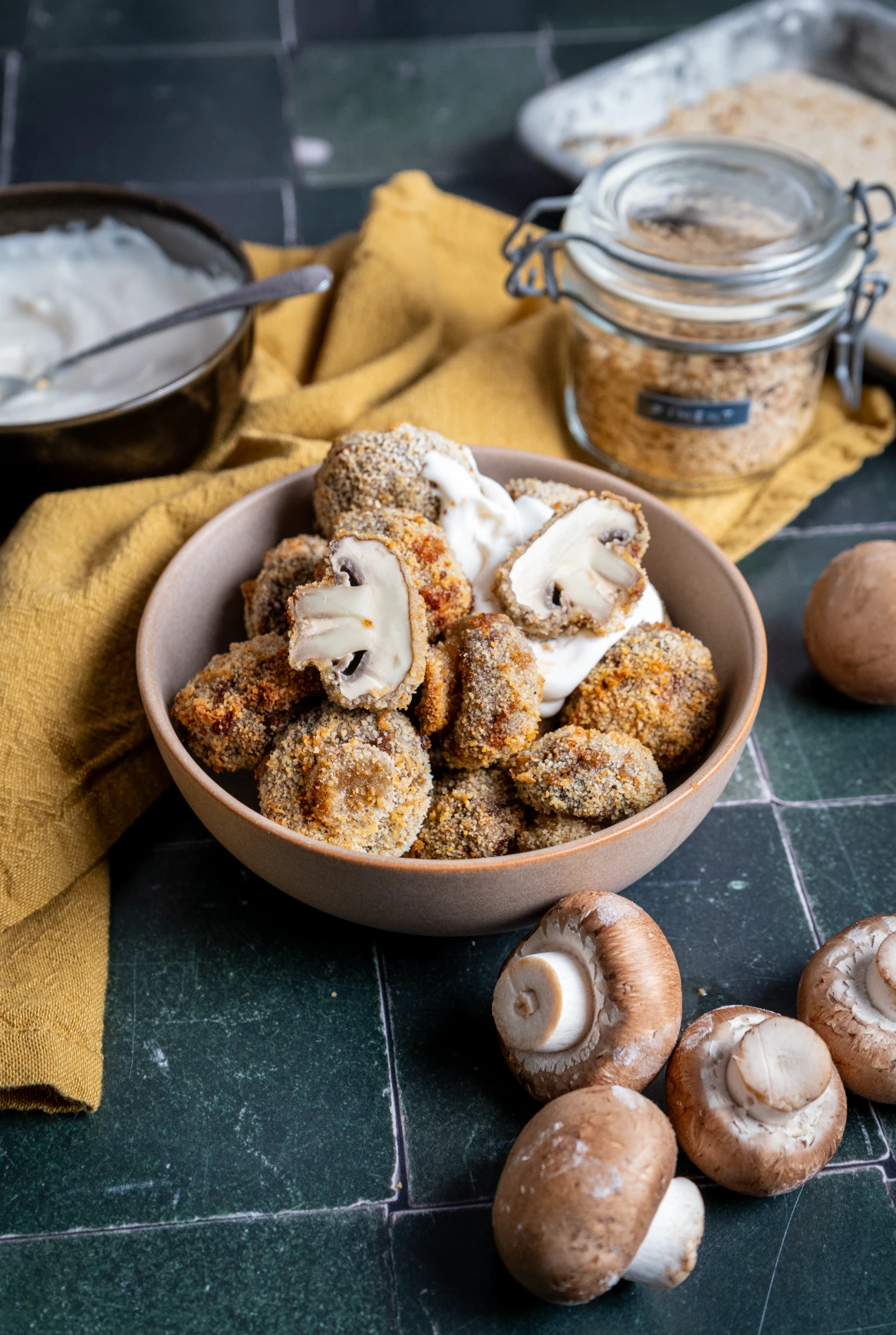 Schüssel mit panierten Champignons und Dip, umgeben von frischen Pilzen und Zutaten auf einem dunklen Tisch.