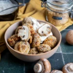 Schüssel mit panierten Champignons und Dip; daneben frische Champignons und Brotkrumen im Glas auf grünem Untergrund.