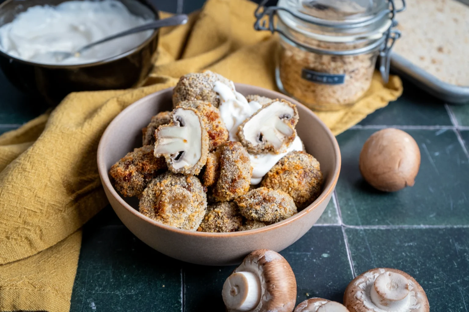 Schüssel mit panierten Champignons und Dip; daneben frische Champignons und Brotkrumen im Glas auf grünem Untergrund.