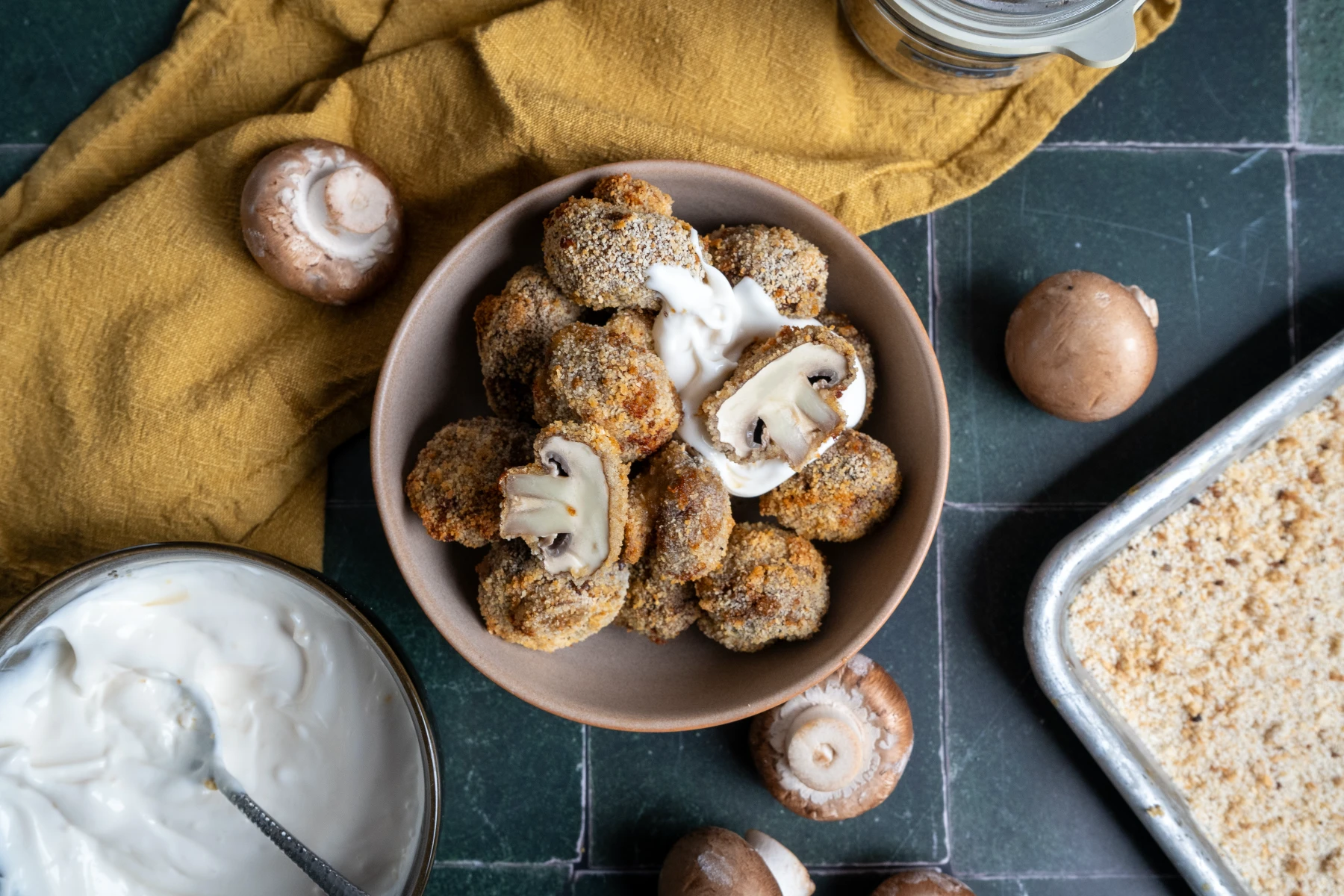 Gebackene, panierte Champignons in einer Schüssel mit Sauerrahm, umgeben von rohen Pilzen und Paniermehl.