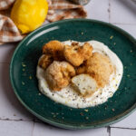 Panierter Blumenkohl auf Joghurtsoße auf einem grünen Teller, daneben eine Zitrone und karierte Serviette.