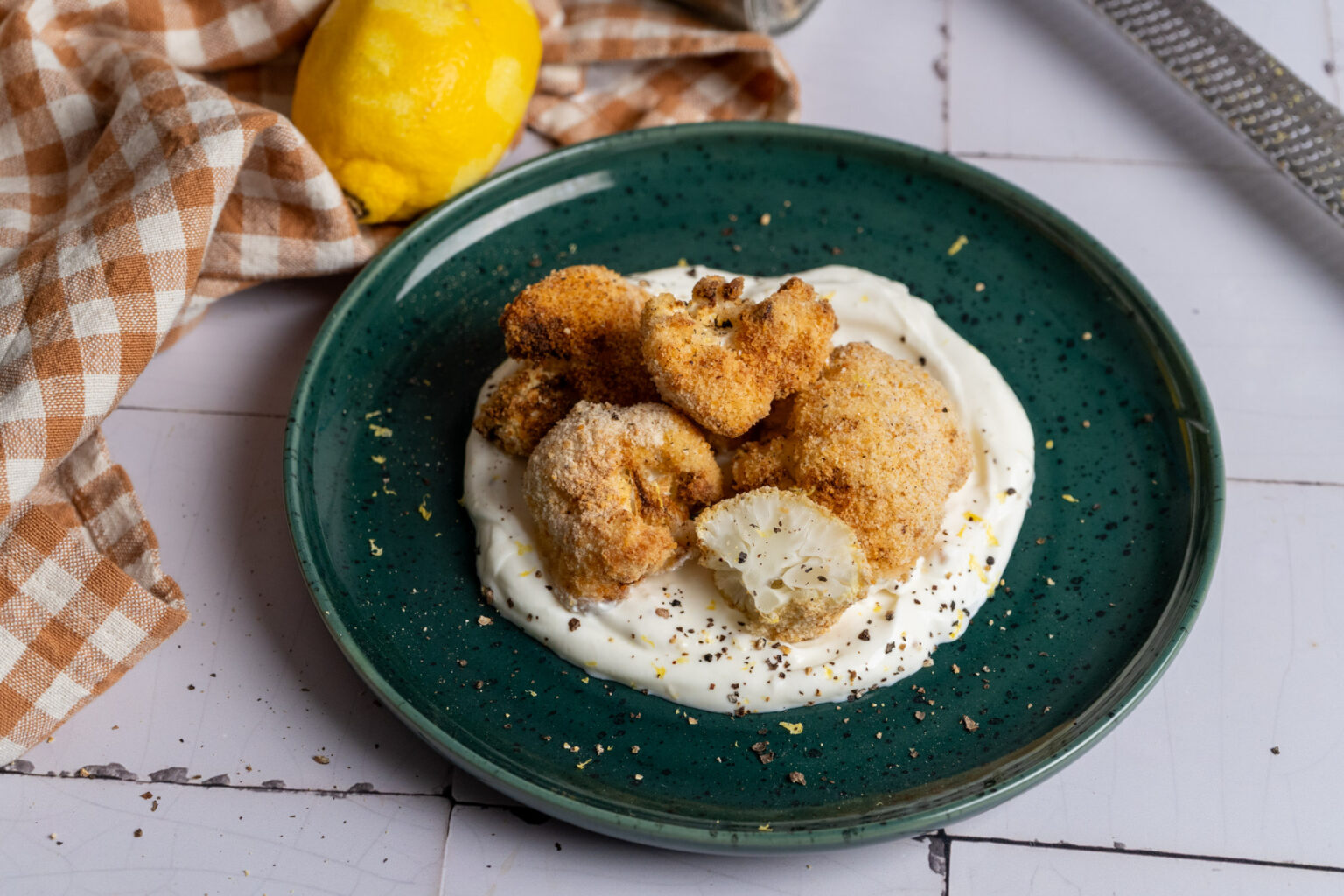 Panierter Blumenkohl auf Joghurtsoße auf einem grünen Teller, daneben eine Zitrone und karierte Serviette.