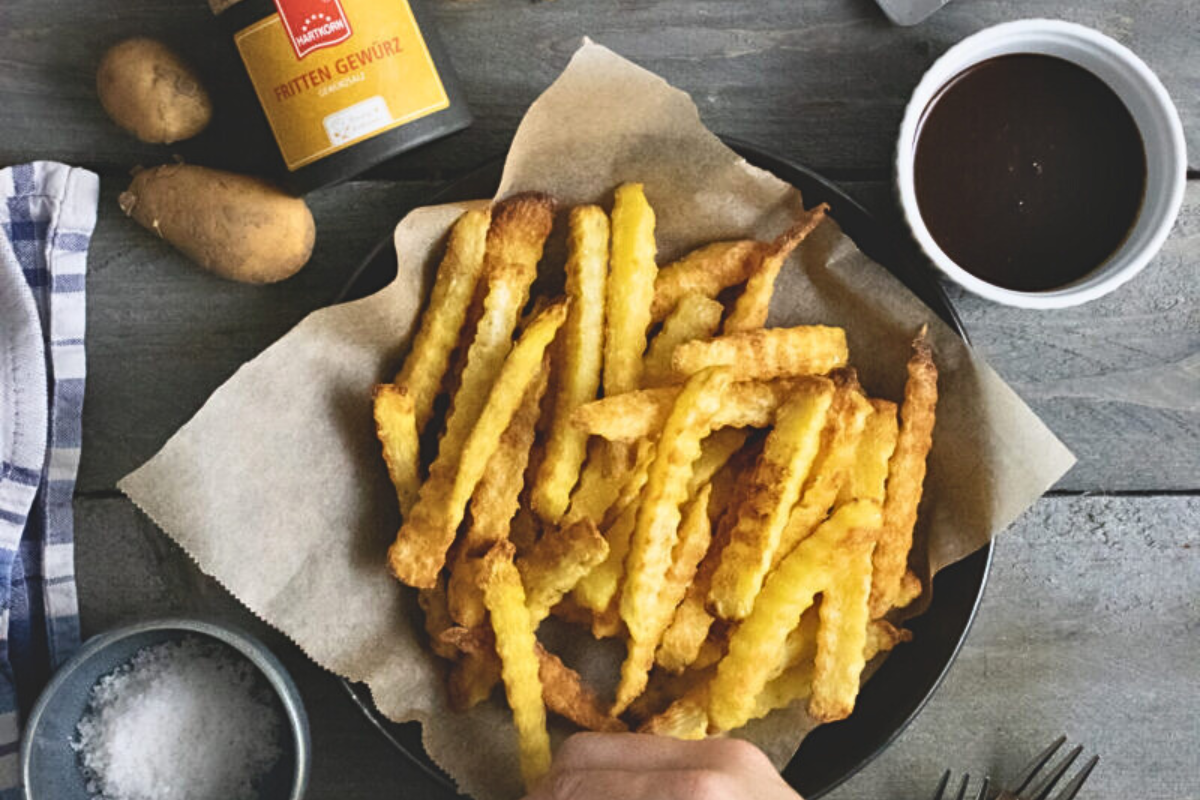 Ein Teller mit gewellten Pommes frites, Pommessauce, Salz, zwei Kartoffeln und einer Gewürzdose auf einem Holztisch.