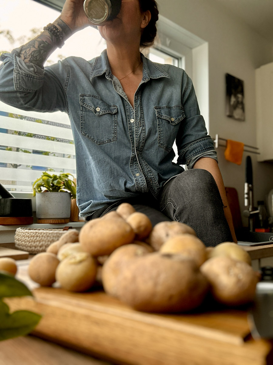 Person in Jeanshemd trinkt aus einer Tasse, sitzt neben einem Haufen Kartoffeln in gemütlicher Küchenszene.
