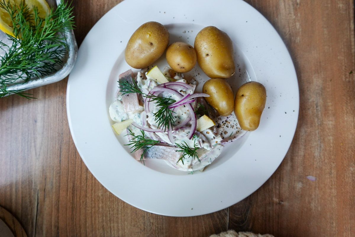 Teller mit Hering in Sahnesauce, roten Zwiebeln und Dill, serviert mit kleinen Kartoffeln auf einem Holztisch.