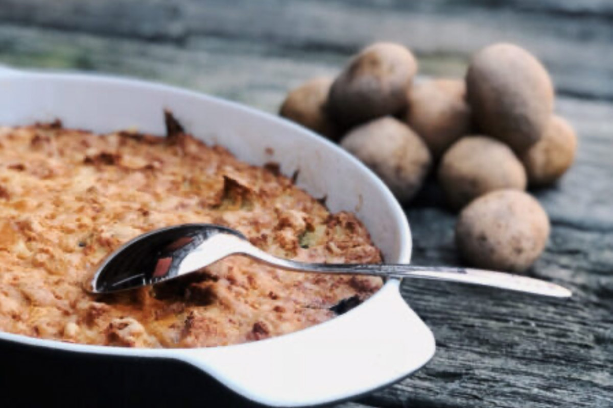 Auflaufform mit Kartoffelauflauf und Löffel, daneben rohe Kartoffeln auf Holztisch.