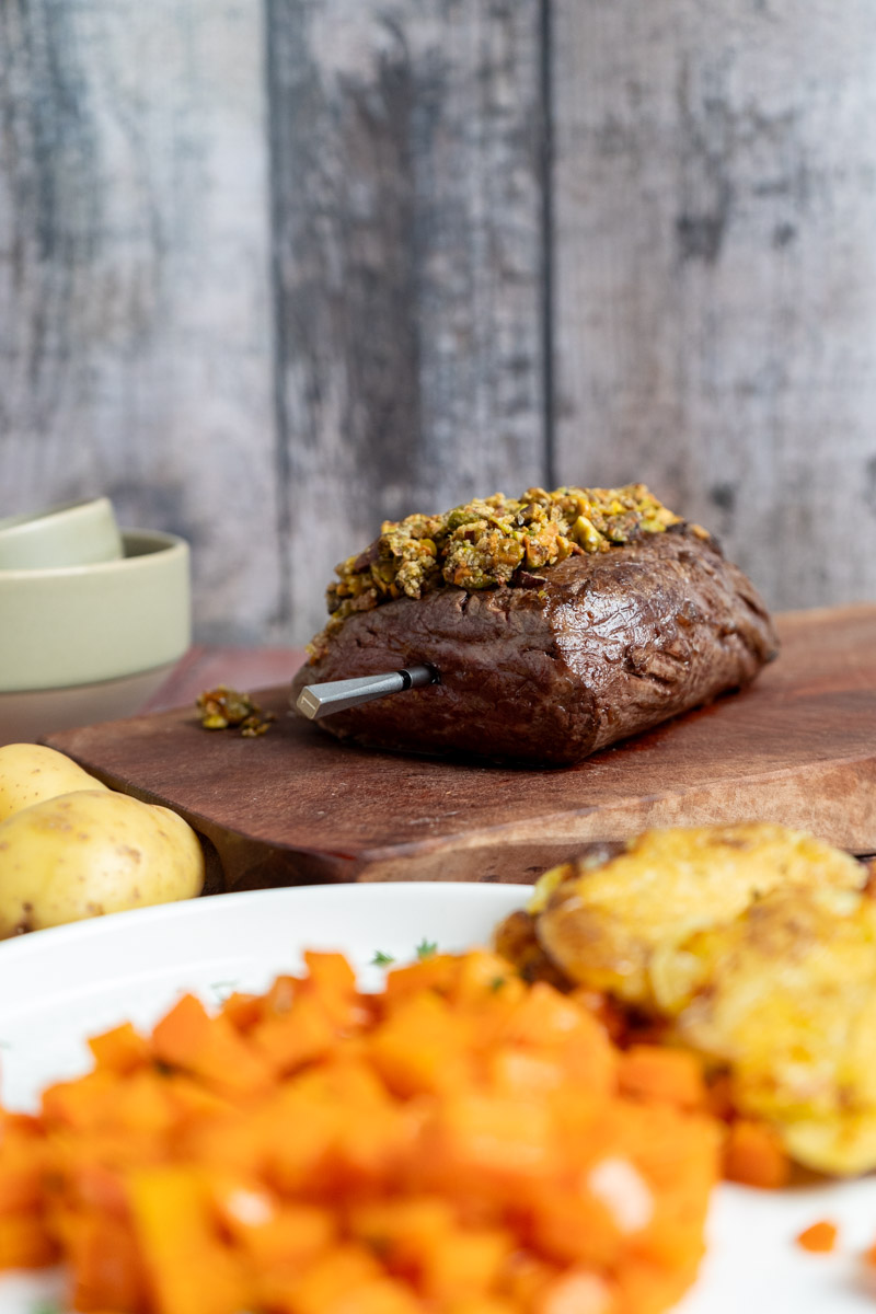 Rinderfilet-2 Braten mit Kräuterkruste auf Holzbrett, vorne Kartoffeln und Karotten auf Teller. Thermometer im Braten.