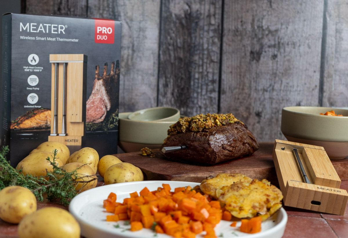 Fleisch mit Drahtthermometer, umgeben von Kartoffeln, Möhren und Kochzubehör auf Holzuntergrund. Verpackung im Hintergrund.