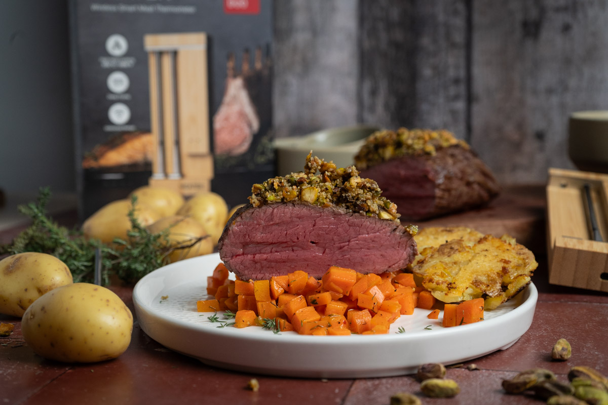 Rinderfilet-05 Ein Teller mit gebratenem Fleisch, gehackten Karotten, Kartoffeln und Nüssen. Dekoration und Verpackung sind im Hintergrund.