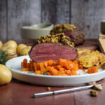 Nahaufnahme eines aufgeschnittenen Rinderbratens mit Kruste, serviert auf Karottenwürfeln und Kartoffeln auf einem weißen Teller.