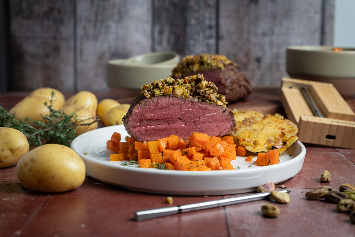 Rosa-gebratenes-Rinderfilet-01 Nahaufnahme eines aufgeschnittenen Rinderbratens mit Kruste, serviert auf Karottenwürfeln und Kartoffeln auf einem weißen Teller.