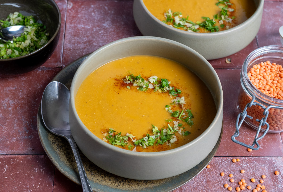 Türkische-Linsensuppe-01 Rote Linsensuppe in einer Schüssel, garniert mit frischen Kräutern und Paprikapulver. Glas mit roten Linsen daneben.