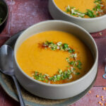Schale mit Linsensuppe, garniert mit frischen Kräutern und Gewürzen, daneben Glas mit roten Linsen und Löffel auf Teller.