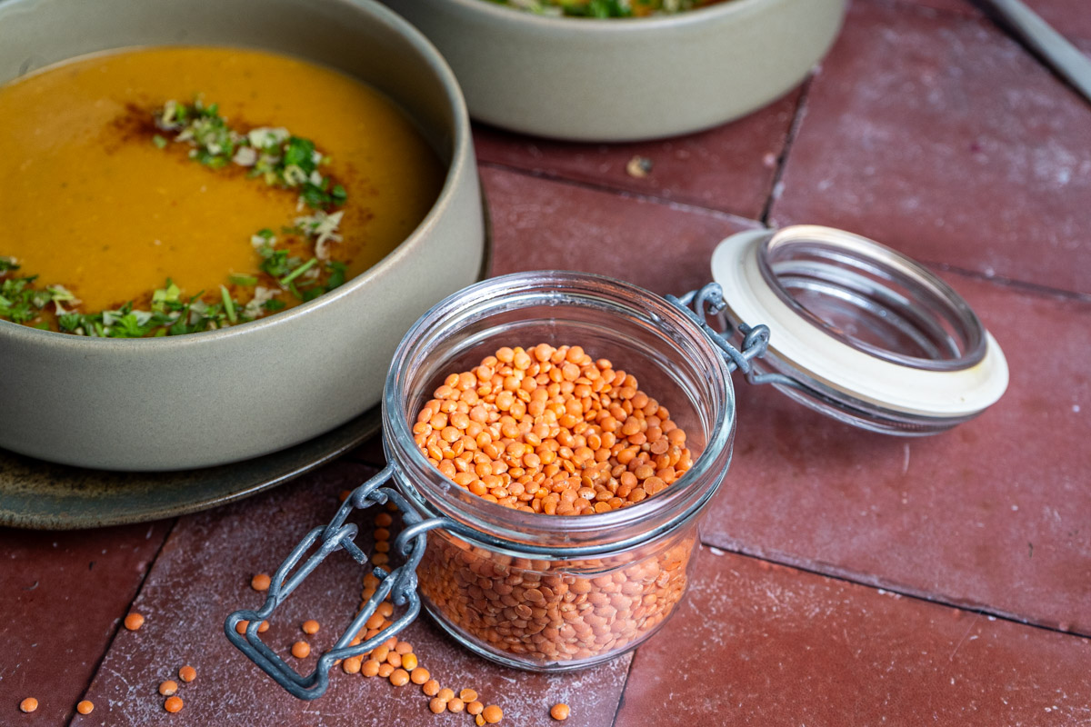 Türkische-Linsensuppe-02 Schüssel mit Linsensuppe, garniert mit Kräutern, neben offenem Glas mit roten Linsen auf rotem Fliesenboden.