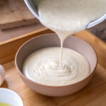 Flüssiger Teig wird aus einem Topf in eine Schüssel gegossen. Daneben stehen aufgeschlagene Eischalen auf einem Holztablett.